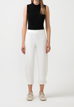 Frau trägt ärmelloses schwarzes Top, weiße kurze weite Hosen und beige Turnschuhe, steht vor schlichtem weißem Hintergrund.