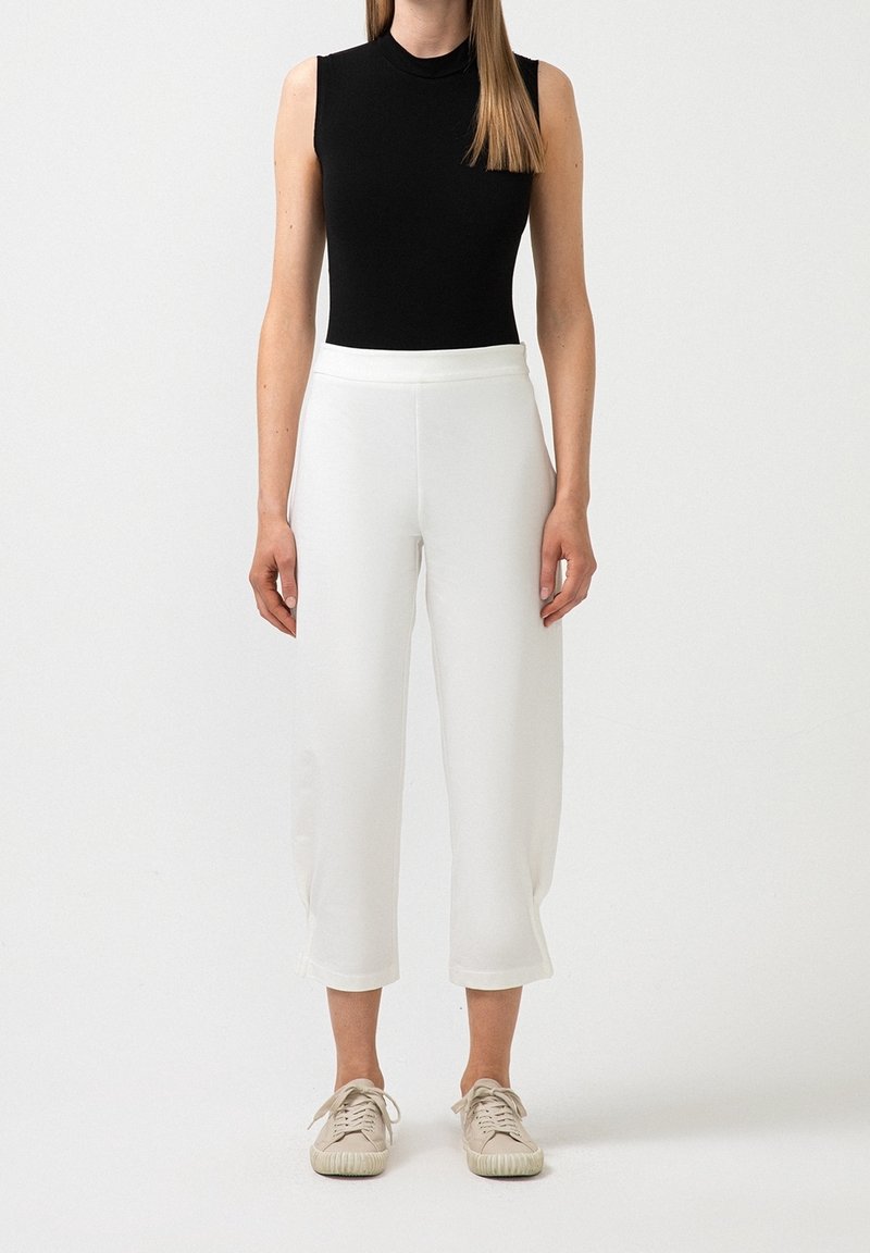 Femme portant un top noir sans manches, un pantalon large blanc raccourci et des baskets beiges, debout devant un fond blanc uni.