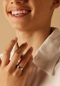 Zilveren metalen ring met een gepolijste afwerking en een horizontale groefdetail, gedragen op een hand met een natuurlijke huidskleur, tegen een beige achtergrond.