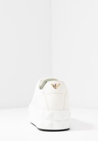 Valkoiset sneakerit, joissa on teksturoitu, geometristi muotoiltu kumipohja. Yläosa on sileää nahkaa ja kantapäässä on kultainen logo. Minimalistinen muotoilu.