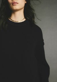Pulover negru tricotat cu un gât înalt și textură cu nervuri. Asortat cu un colier din lanț aurit cu zale alungite.