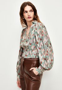 Blusa a fantasia verde con grandi punti colorati; presenta maniche lunghe a sbuffo e scollatura a V. Abbinata a pantaloni in pelle marroni.