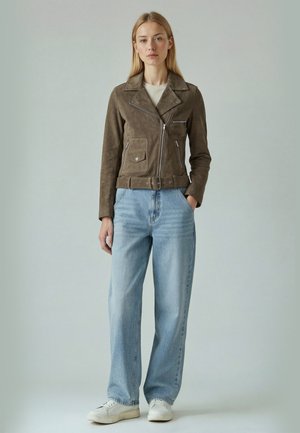 Fiatal nő barna bőr motoros dzsekiben, világoskék, bő szárú farmernadrágban és fehér tornacipőben, kezét zsebében tartva áll.