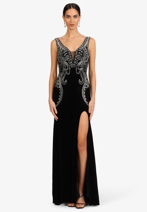 Frau trägt ein schwarzes ärmelloses Kleid mit silbernen Perlendetails, tiefem Ausschnitt, hohem Frontschlitz und schwarzen Riemchenabsätzen.