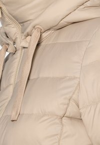 Beżowa pikowana kurtka z wysokim kołnierzem, regulowanym ściągaczem i teksturowaną tkaniną z pikowanym przeszyciem, zapinana na zamek błyskawiczny z przodu.