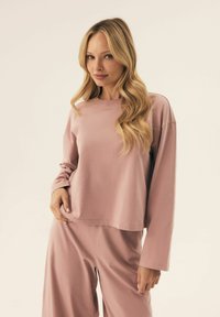 Jasnoróżowa, długiego rękawa bluzka z okrągłym dekoltem i subtelnym napisem na klatce piersiowej, w zestawie z pasującymi spodniami o szerokich nogawkach. Miękka tekstura.