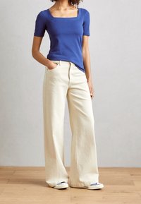 Blaues T-Shirt mit quadratischem Ausschnitt und kurzen Ärmeln, kombiniert mit weit geschnittenen, cremefarbenen Hosen, die eine glatte Textur und einen lässigen Sitz bieten. Weiße Schuhe sind enthalten.