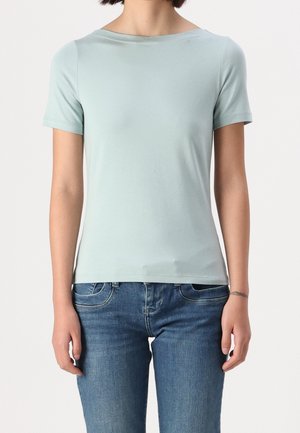 Femme portant un t-shirt bleu clair uni à manches courtes et un jean bleu, debout avec les bras détendus le long du corps devant un fond uni.