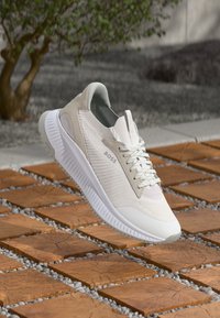 Beige and white BOSS sneaker with knit upper and thick sole floating above wooden tiles with small gravel between them outdoors.