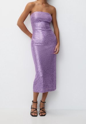 Mujer con un vestido midi sin tirantes, ajustado y de lentejuelas lavanda, y sandalias negras de tacón alto con tiras, posando frente a una pared blanca lisa.