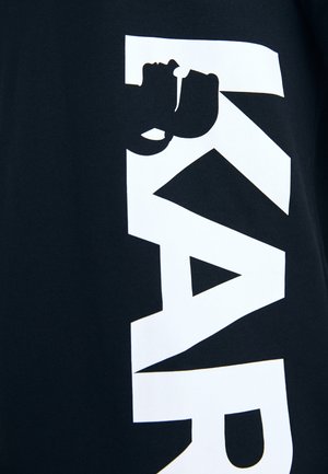 Maglietta nera in cotone con grandi lettere bianche a blocchi e dettaglio sagomato. Il design enfatizza una tipografia audace ed elementi grafici.