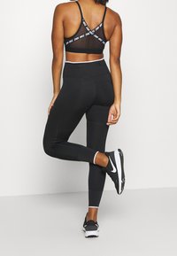 Reggiseno sportivo nero con dettagli in rete e spalline con logo, abbinato a leggings neri a vita alta con accenti testurizzati e scarpe da ginnastica nere.