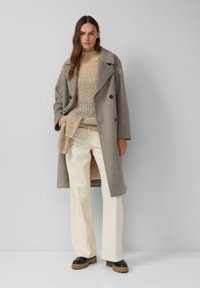 Frau mit langen braunen Haaren, die einen beigen Strickpullover, einen grauen Wollmantel, cremefarbene Hosen und schwarze Stiefel mit braunen Sohlen trägt, steht vor einer grauen Wand.