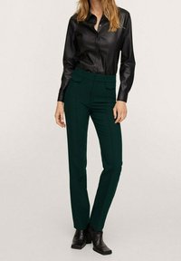 Chemise noire à boutons en simili cuir, pantalon tailleur vert avec poches latérales, et bottes noires à talons. Texture lisse, silhouette ajustée.