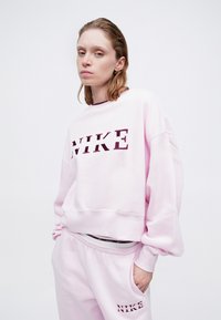 Ružový mikina a tepláky s voľným strihom. Mikina má výrazné písmená "NIKE" v tmavšej farbe. Hladká textúra látky.