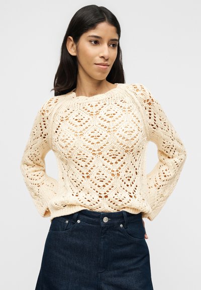 Γυναίκα που φοράει πλεκτό πουλόβερ σε κρεμ απόχρωση με διαμαντένια σχέδια και σκο ύρα μπλε τζιν με ψηλή μέση, ποζάροντας με τα χέρια πίσω από την πλάτη της.