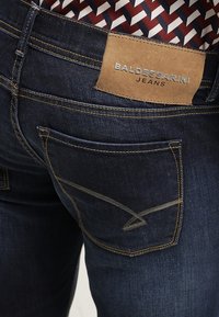 Mörka denimjeans med en läderetikett märkt "BALDESSARINI JEANS." Har kontrasterande guldstickningar och vinklade bakfickor.