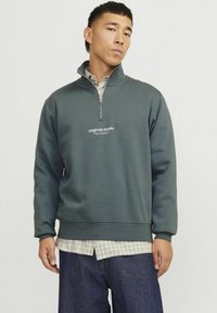 Mørkegrøn fleece pullover med kvart lynlås og et logo på brystet, lagdelt over en ternet skjorte, og i kombination med mørke denim jeans.