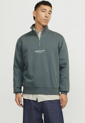 Sudadera de forro polar de color verde oscuro con cremallera de cuarto, con un logo en el pecho, superpuesta a una camisa a cuadros, combinada con jeans de mezclilla oscura.