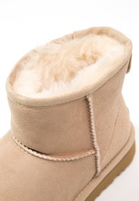 Botines de ante beige con forro de piel de terciopelo crema, costuras visibles y suela de goma. El puño es suave y se pliega hacia afuera.