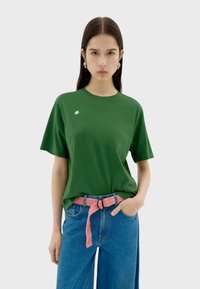 Groene T-shirt met korte mouwen en een ronde halslijn, voorzien van een klein wit logo op de linkerkant van de borst, gecombineerd met high-waisted blauwe jeans en een roze riem.