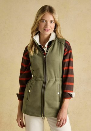 Femeie blondă purtând o vestă oliv cu fermoar peste o bluză cu mâneci lungi în dungi maro și roșii și pantaloni albi, stând în picioare pe un fundal bej.