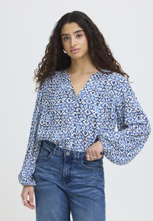 Donna con capelli lunghi e ricci, che indossa una blusa con fantasia blu e bianca e jeans blu a vita alta, in piedi con una mano sulla vita.