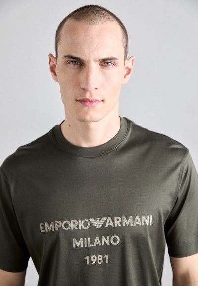 Oliivroheline puuvillane T-särk, millel on heledat halli värvi tekst "EMPORIO ARMANI MILANO 1981", ümmargune kaelus ja lühikesed varrukad.