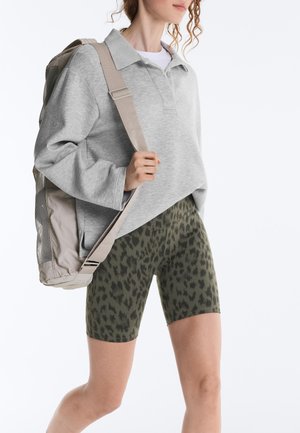 Persoon die een lichtgrijs overhemd met lange mouwen en kraag draagt, olijfgroene biker shorts met patroon en een beige rugzak over één schouder draagt.