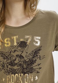 Camiseta de algodón verde oliva con cuello redondo, que presenta gráficos de un águila negra y texto en foil dorado que dice "EST-75 ROCK ON."