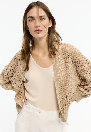 Junge Frau trägt eine beige offene Strickjacke über einem cremefarbenen Tanktop und weißen Hosen mit hoher Taille, steht mit den Händen in den Hüften.