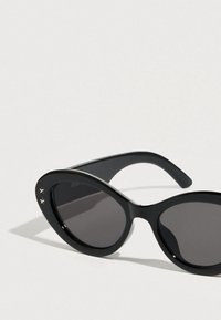 Lunettes de soleil noires à monture œil de chat avec verres gris, finition brillante et deux embellissements en forme d'étoile sur la monture. Design incurvé et branches flexibles.