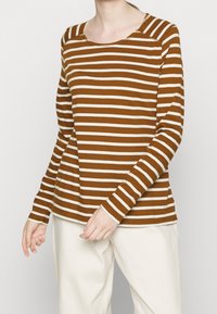 Brown and cream striped long-sleeve shirt with a round neckline and visible stitching detail, paired with light-colored pants.