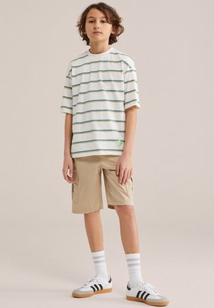 Niño de pie con camiseta blanca a rayas, pantalones cortos cargo beige, calcetines blancos y zapatillas blancas con rayas negras sobre fondo liso.