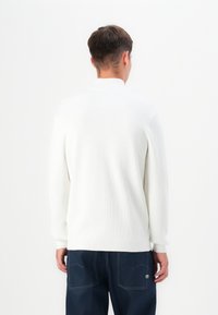 Suéter blanco de canalé con un alto cuello y mangas largas, combinado con pantalones azul oscuro que presentan costuras visibles y una etiqueta en el bolsillo.
