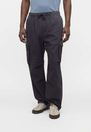 Homme portant un pantalon cargo noir ample avec des poches latérales et des baskets beiges, debout sur un fond blanc uni.
