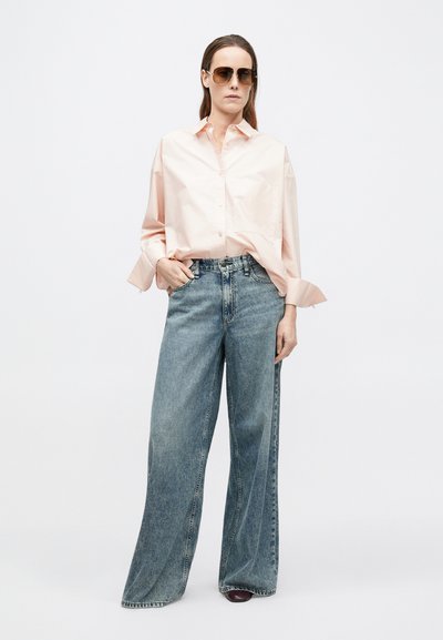 Hellrosa übergroßes Hemd mit aufgerollten Manschetten, kombiniert mit weit geschnittenen hellblauen Jeans. Model trägt dunkle Sonnenbrille.