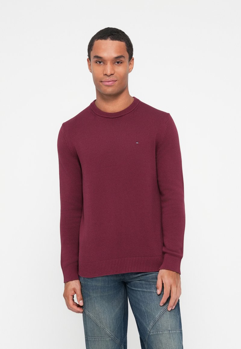 Bordo pleten pulover z okroglim izrezom in rebrastimi manšetami. V kombinaciji z jeans hlačami z diskretnimi šivi. Preprost logotip na prsih.
