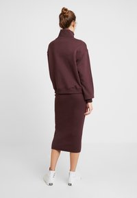 Mujer con el cabello recogido en un moño, vistiendo un sudadera burdeos de cuello alto y una falda midi a juego, de pie, de espaldas, con zapatillas blancas de caña alta.