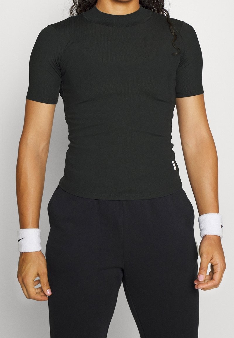 Top negro de manga corta ajustado hecho de tela suave y elástica con cuello alto. Se lleva con pantalones negros y muñequeras blancas.