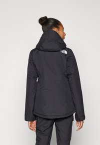 Schwarze, isolierte Jacke mit Kapuze, glatter Textur und Logo auf der linken Schulter. Verfügt über einen längeren Rücken und elastische Bündchen.