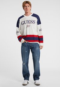 Langärmliges weißes Hemd mit horizontalen Streifen in Marineblau und Rot, "GUESS" in großen Buchstaben aufgedruckt. Getragen mit verwaschenen blauen Jeans und weißen Sneakers.