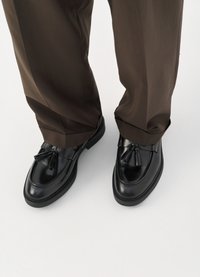 Black leather loafers with a rounded toe, decorative tassels, and textured stitching, paired with brown, slightly loose trousers.