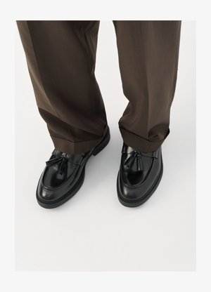 Black leather loafers with a rounded toe, decorative tassels, and textured stitching, paired with brown, slightly loose trousers.