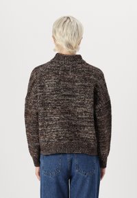 Braun-grauer grob gestrickter Pullover mit hohem Kragen, überschnittenen Schultern und gerippten Bündchen, kombiniert mit blauen, hoch taillierten Jeans.