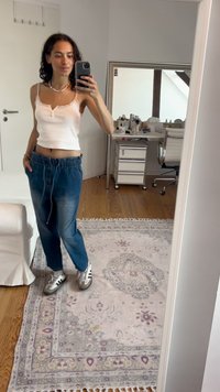 Hvit ermeles topp med blondekant, blå avsmalnende denimbukser med elastisk midjebånd, og grå sneakers med svarte striper. Tregulv og mønstret teppe.