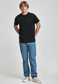 Czarny t-shirt z krótkim rękawem z dyskretnym logo na piersi, w połączeniu z jasnoniebieskimi dżinsami i beżowymi trampkami, na jasnym tle.