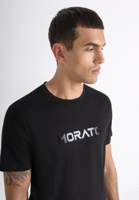 Camiseta de algodón negra con un logotipo metálico plateado "MORATO" en el pecho. Cuello redondo, mangas cortas y un diseño ajustado.