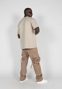 Beige bomulds t-shirt og lysebrune cargo bukser med flere lommer. Hvide sneakers. Model med ryggen til, solbriller, tekstureret stof.