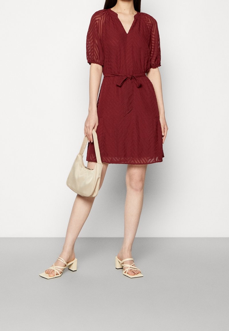 Femme portant une robe bordeaux mi-longue à manches bouffantes, tenant un sac à main beige et portant des sandales crème à talons blocs et lanières.
